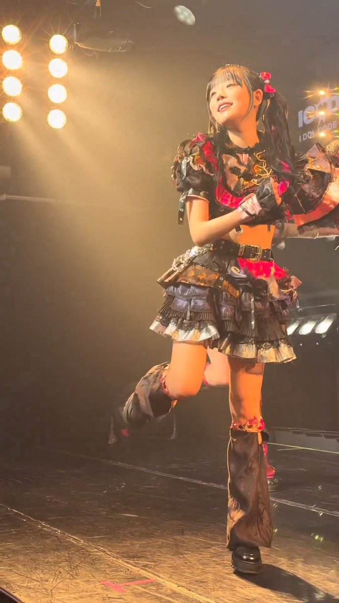 A female performer with long dark hair in pigtails performs on stage wearing a black and red outfit with a ruffled skirt, dynamic pose with one leg raised under bright spotlights.