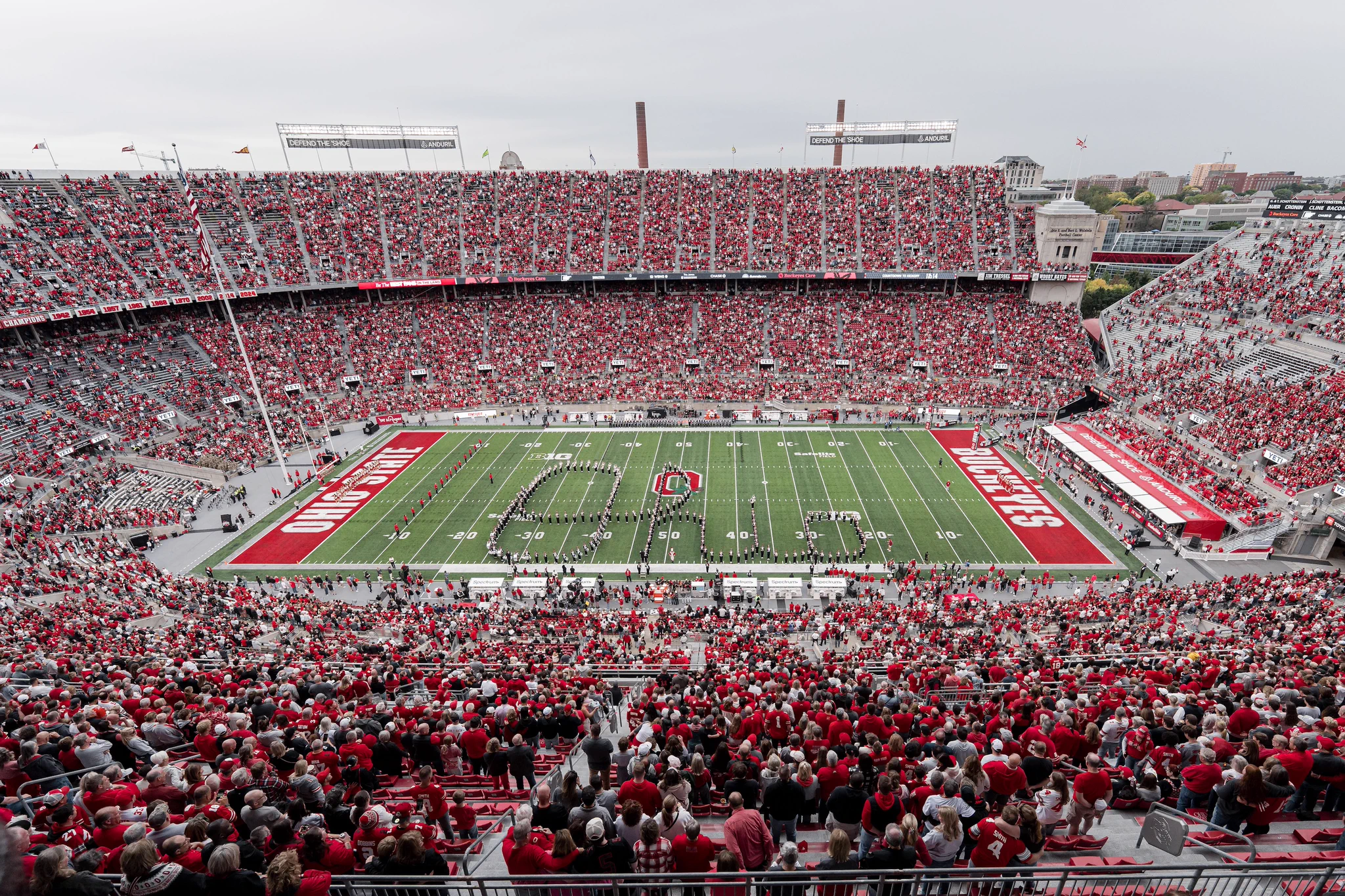 A large football stadium filled with spectators, predominantly wearing red clothing. The field features "Ohio State" and the Ohio State logo at the center. The stands are packed, and the atmosphere is lively.
