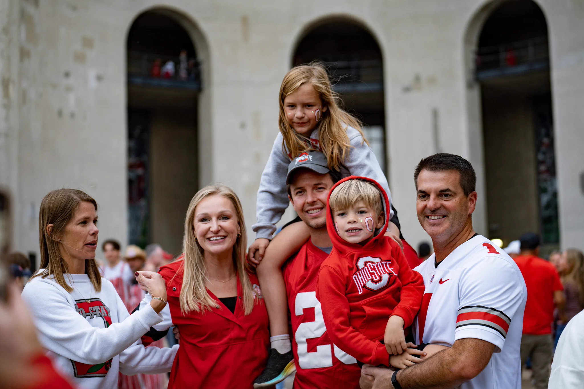A group of people wearing red Ohio State attire stand in front of large arches at a stadium. One person carries a child on the shoulders, while another holds a smiling child. The atmosphere appears joyful and celebratory.