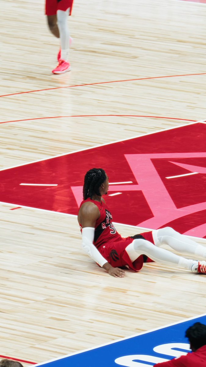 A basketball player in a red Rockets jersey sits on the court floor near a large red star logo, appearing to recover from a play.