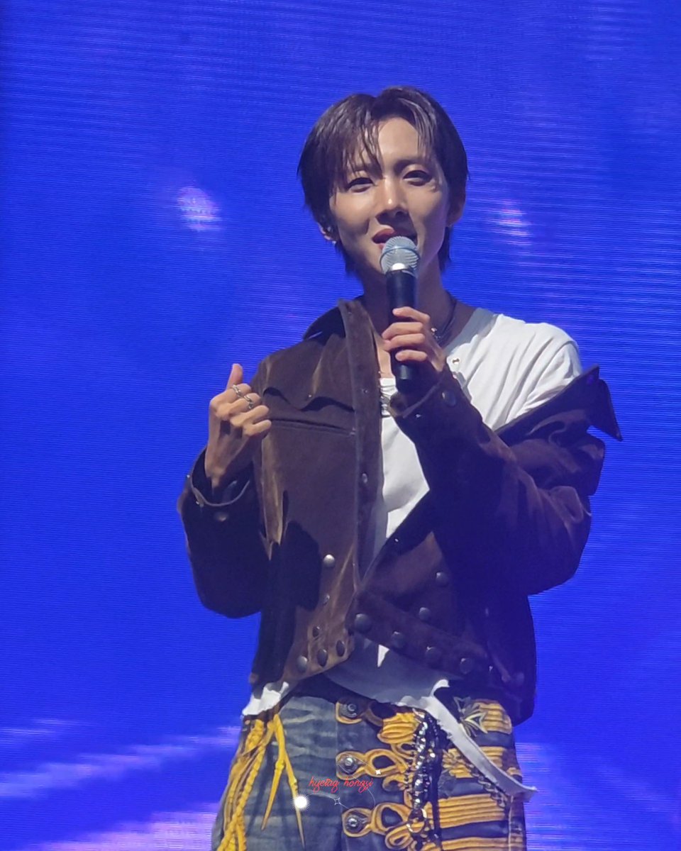 A young male performer with short brown hair sings into a microphone on stage, wearing a brown jacket over a white shirt with patterned pants.