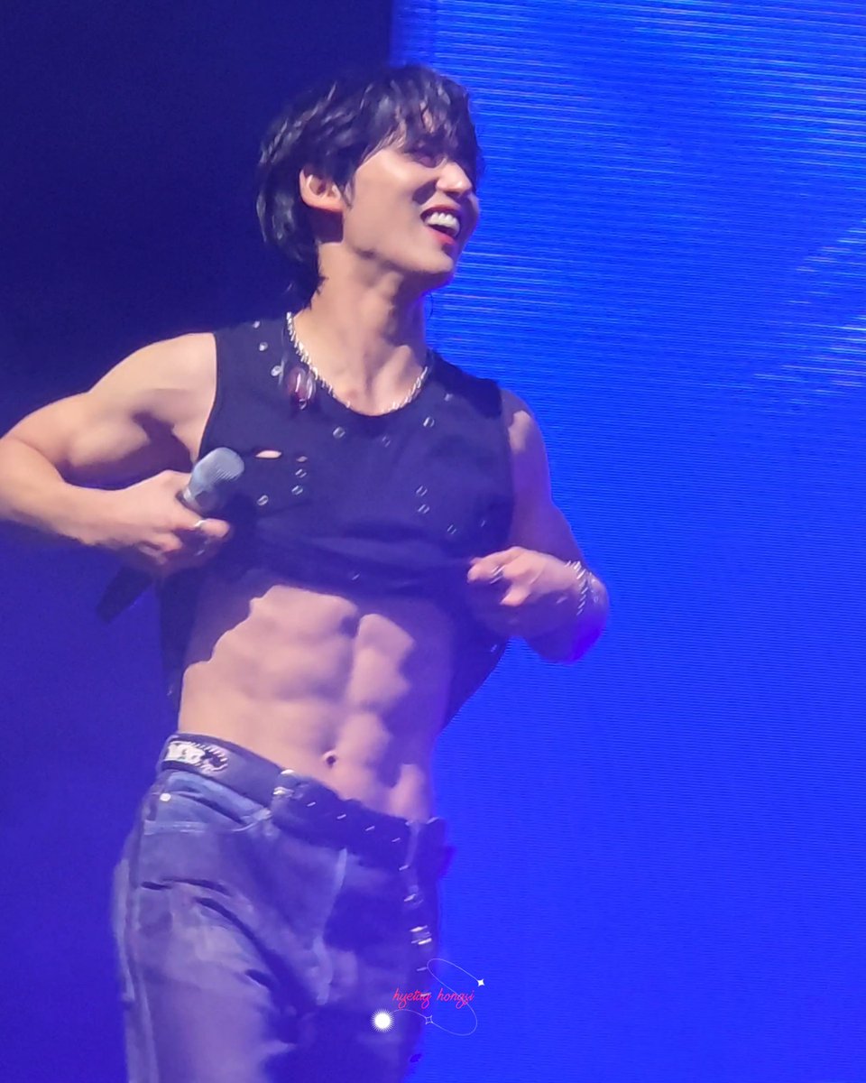A performer in a black studded cropped tank top lifts his shirt to reveal defined abs while smiling on stage with a blue-lit background; a watermark reads 'krystal honggi'.