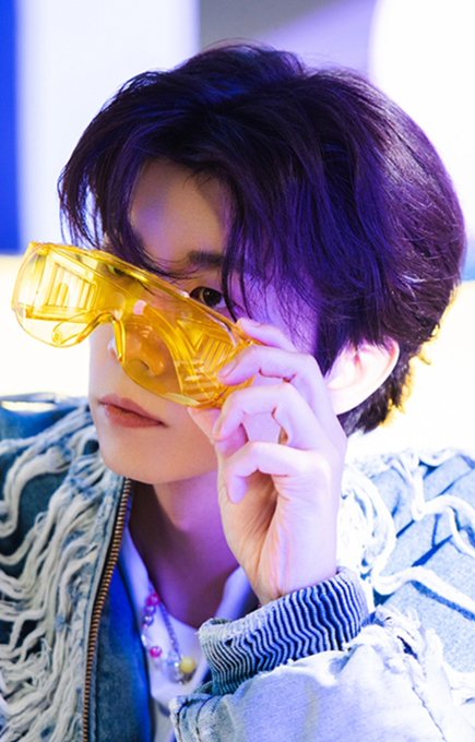 A close-up portrait of a young person with dark purple-tinted hair, wearing yellow translucent sunglasses and a distressed denim jacket with fringe details.