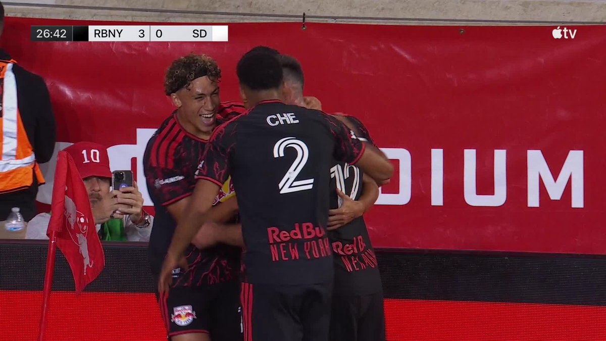 A broadcast screenshot from Apple TV showing New York Red Bulls soccer players celebrating a goal; the scoreboard reads '26:42 RBNY 3 0 SD' while players wearing black and red jerseys with 'CHE 2' visible hug each other in front of a red stadium backdrop.