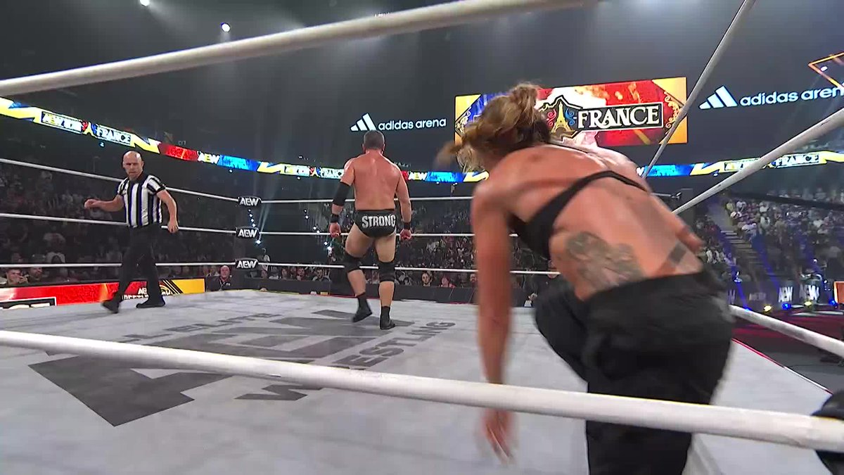 Two female wrestlers face off in a ring marked with "AEW" logos at the Adidas Arena, with the screens behind them displaying "FRANCE." One wrestler with a ponytail and tattoos appears ready to engage a male wrestler wearing "STRONG" trunks, while a referee in stripes looks on, capturing the intense moment from the AEW Collision Grand Slam France event.