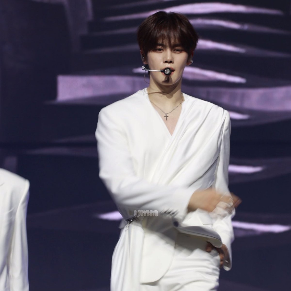 A male performer on stage wearing a flowing white wrap-style outfit with a cross necklace and headset microphone, with 'umm' text visible on his sleeve.