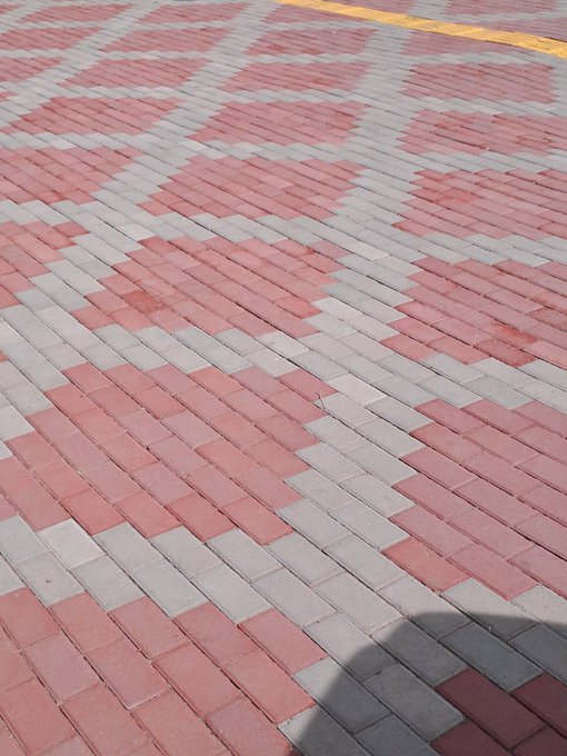 A close-up view of decorative pavement featuring red and gray paving bricks arranged in a geometric pattern, with a partial yellow line and a shadow visible at the bottom.