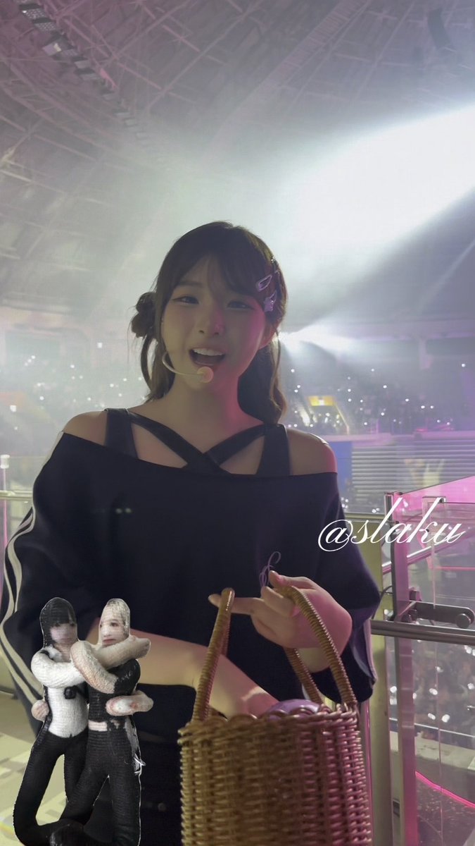 A young woman wearing a headset microphone and a black cutout top smiles at a concert or event, holding a woven basket, with the watermark '@slaku' and an image of two wrestlers visible on the frame.