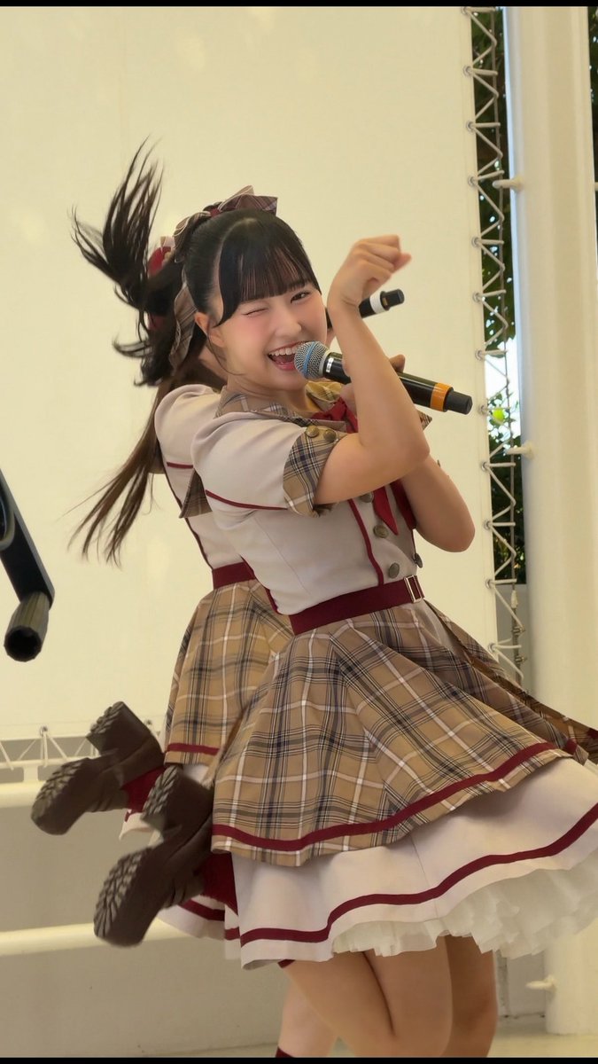A young performer with dark hair in pigtails wearing a plaid school-uniform-style dress winks and smiles broadly while energetically performing on stage with a microphone.
