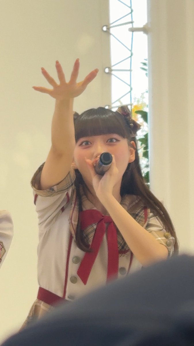 A young woman with long dark hair, wearing a plaid outfit with a red bow, holds a microphone while reaching her hand forward, possibly a member of AKB48.