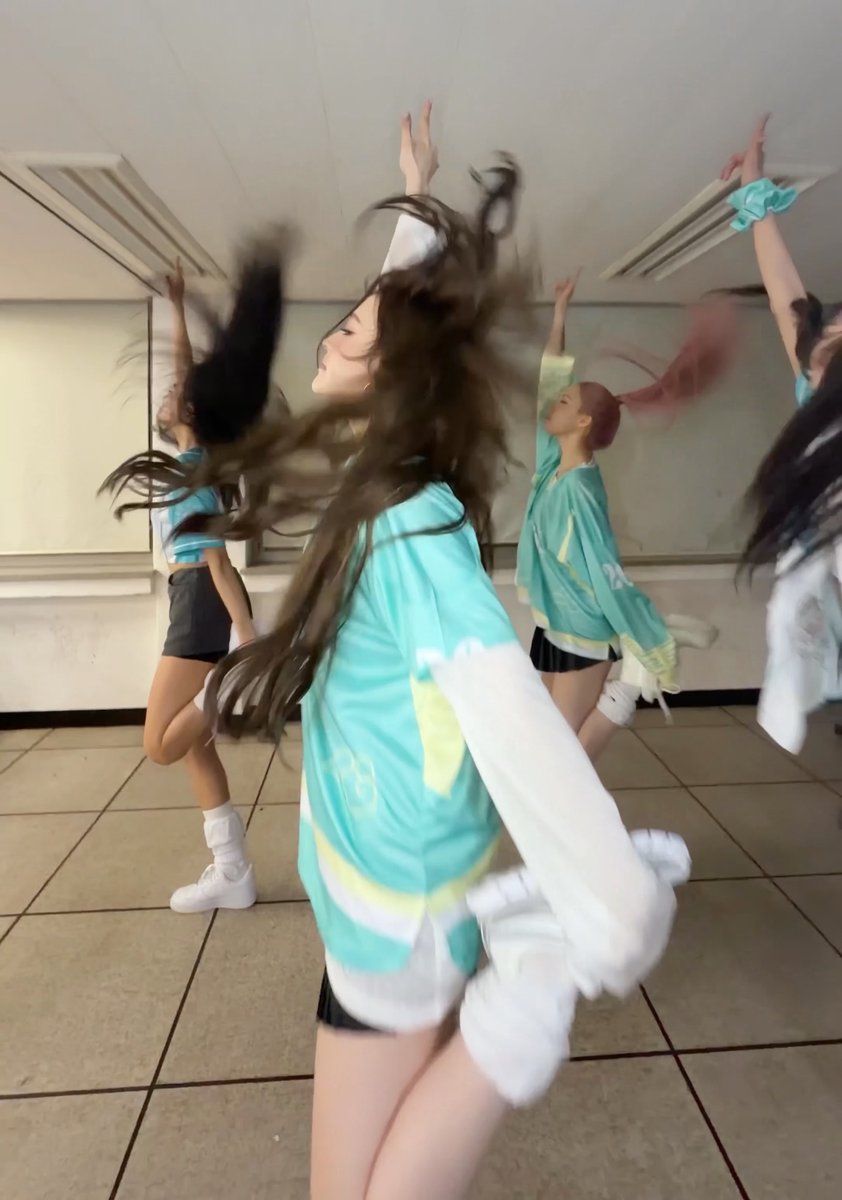 A group of young women in matching teal, white, and black outfits perform a dance routine in a studio, captured mid-motion with their arms raised and hair flying.