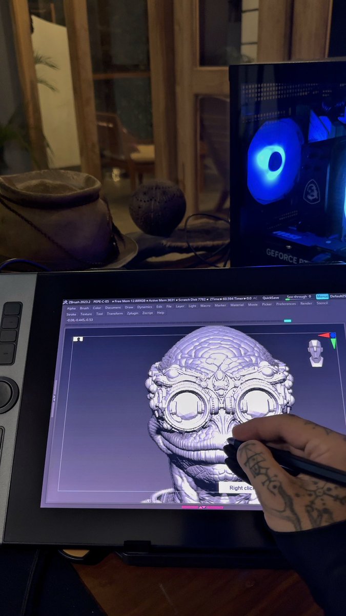 A person's tattooed hand works on a 3D modeling software (ZBrush) displayed on a drawing tablet, with a GeForce RTX PC featuring a glowing blue eye visible in the background.