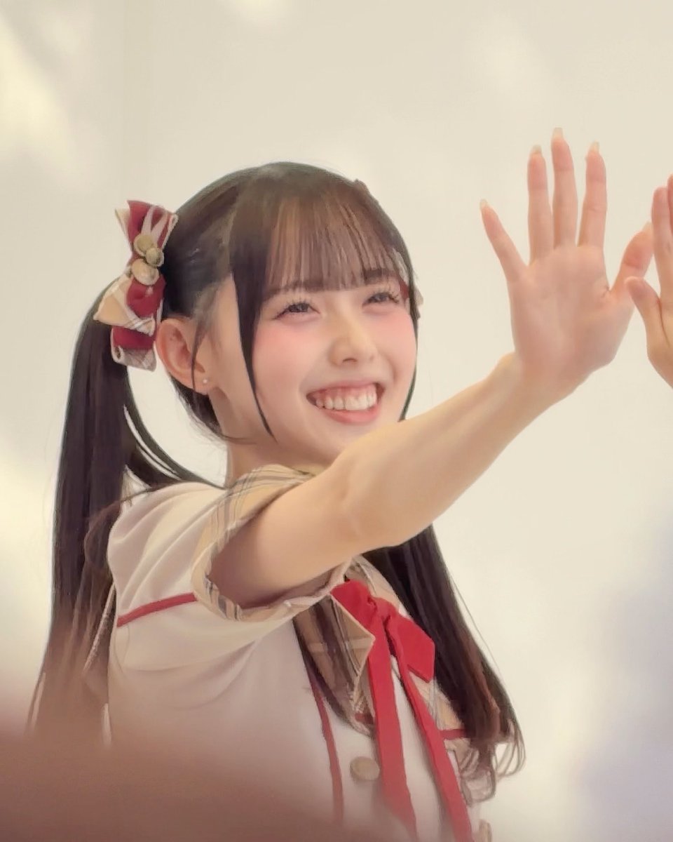 A smiling young woman with dark pigtails and plaid hair bows raises her hand, wearing a white and red plaid outfit.