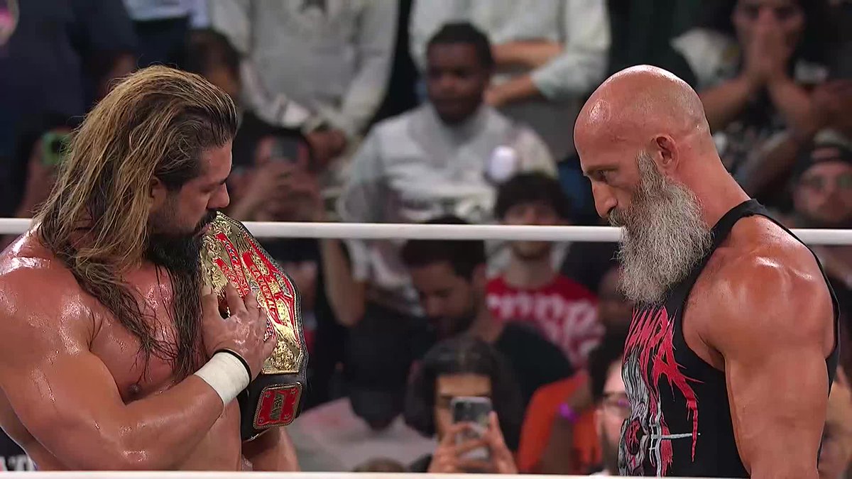 Two muscular professional wrestlers face off in a ring; the wrestler on the left has long hair and holds a gold championship belt, while the bald, bearded wrestler on the right wears a black and red tank top.
