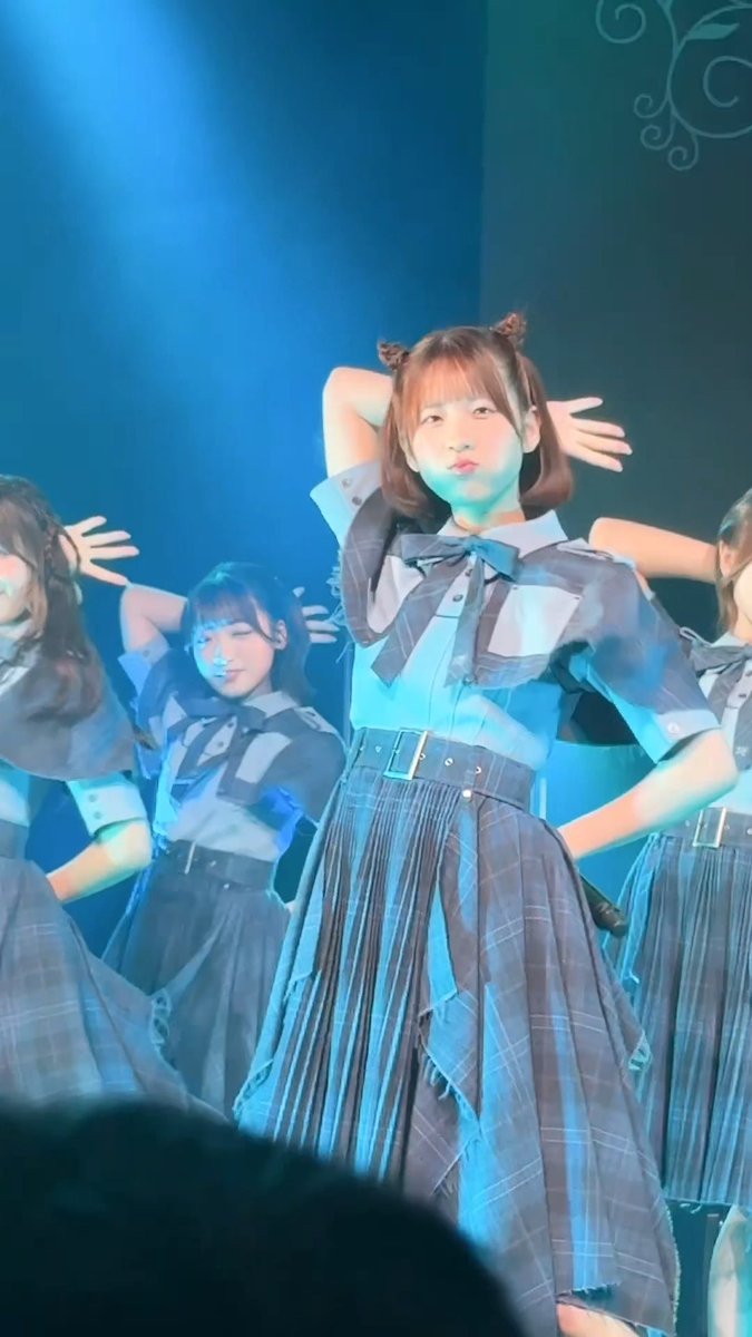 A young performer with twin buns poses on stage in a blue school-uniform-style outfit with a pleated skirt, illuminated by blue stage lighting alongside other performers.