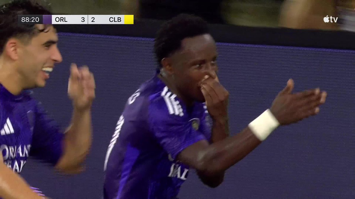 Two Orlando City SC players celebrate on the pitch after Iván Angulo scored to secure their victory over Columbus Crew (ORL 3-2 CLB) at the 88:20 mark of the match. One player covers his face with his hand in an emotional reaction, while his teammate smiles broadly beside him, reflecting the dramatic end-to-end play described in the caption.