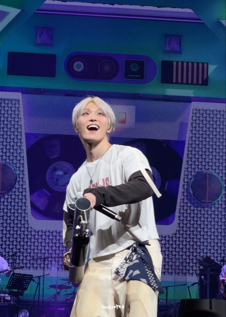 A blonde-haired performer smiles while holding a water bottle on stage, wearing a white t-shirt and beige pants, with a cassette tape-themed backdrop behind him.