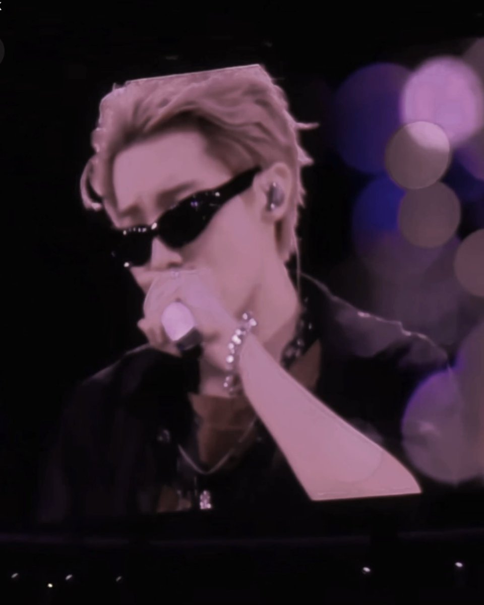 A performer with blonde hair and dark sunglasses sings into a microphone on stage, with pink and purple lighting in the background.