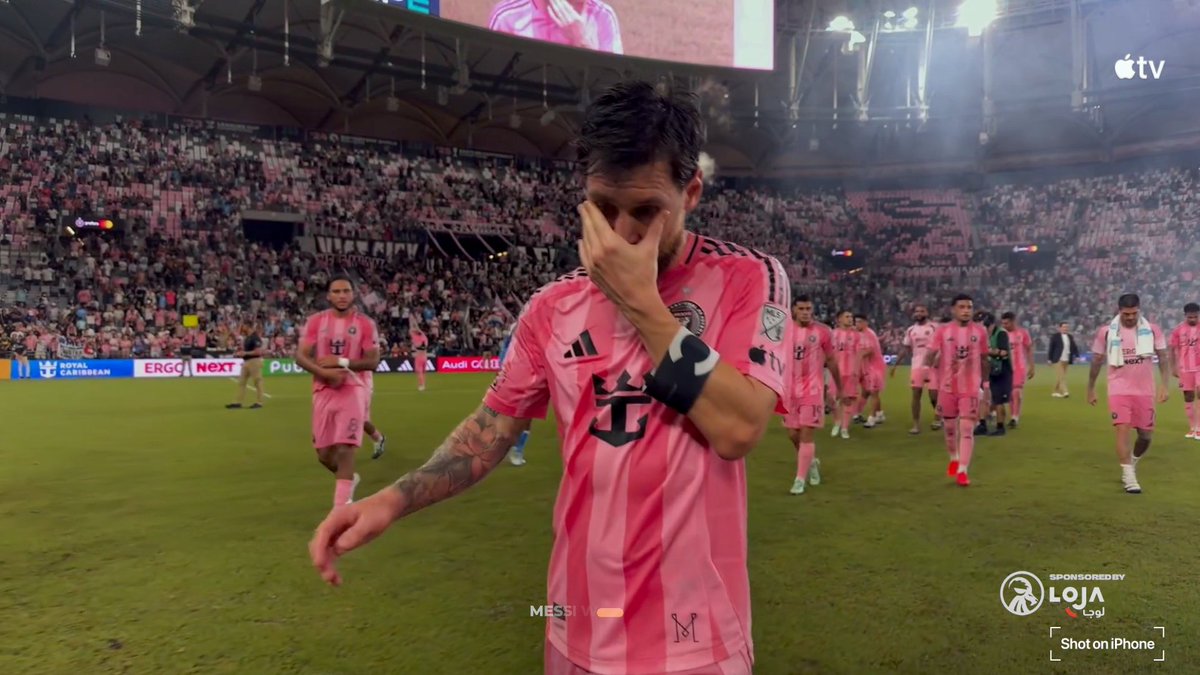 Lionel Messi, wearing the pink Inter Miami kit with the number 10, walks on the field wiping his eye while teammates follow in a stadium setting.