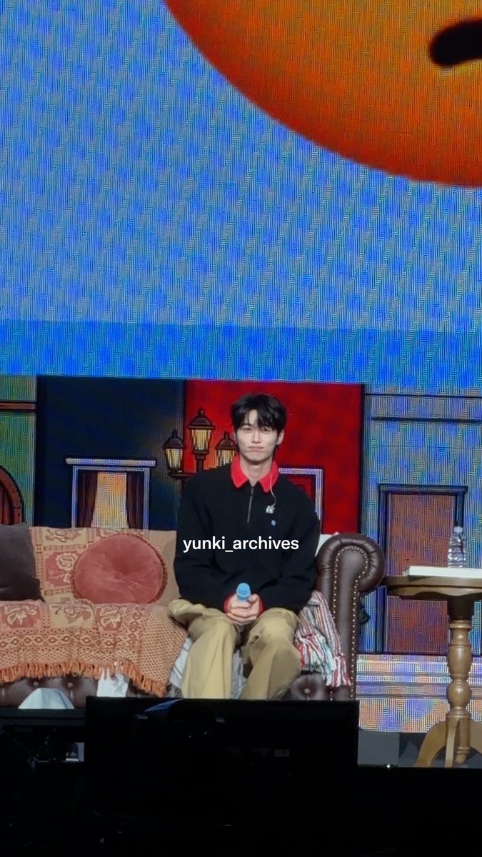 A young man sits on a couch on a stage set, wearing a black sweater over a red collared shirt and tan pants, with 'yunki_archives' watermarked on the image.