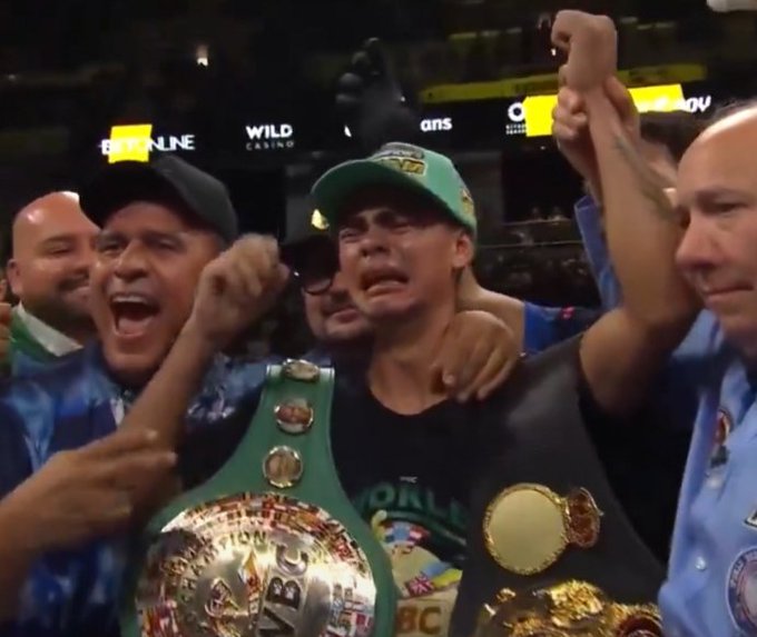 Boxer Canelo Álvarez, wearing a green cap and WBC championship belt, appears emotional with a raised fist while surrounded by a celebrating entourage in an arena setting.