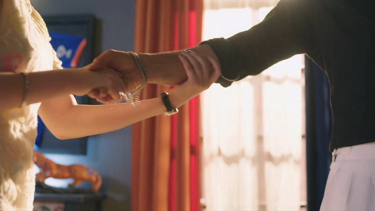 Two people hold hands in a warmly lit indoor setting with red curtains in the background; one person wears a fluffy white garment and the other a dark sweater.