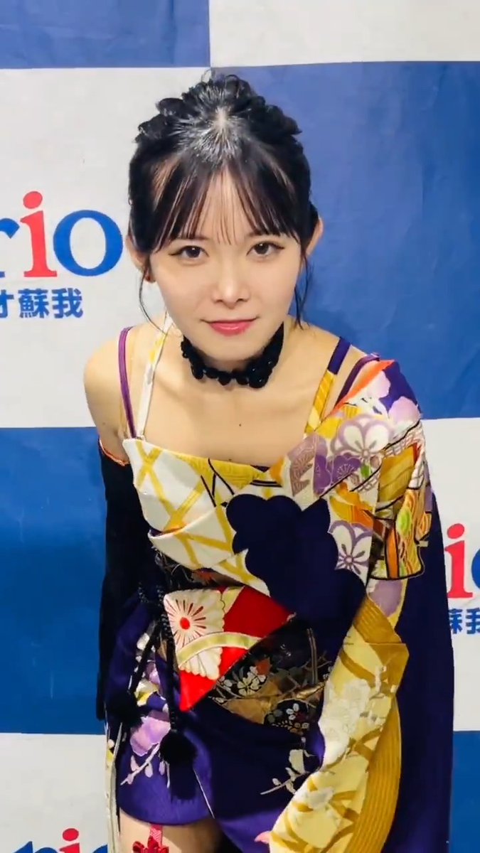 A young woman with dark hair styled up and bangs wears a colorful kimono with floral patterns and a black choker necklace, posing in front of a blue backdrop with partial 'rio' text.