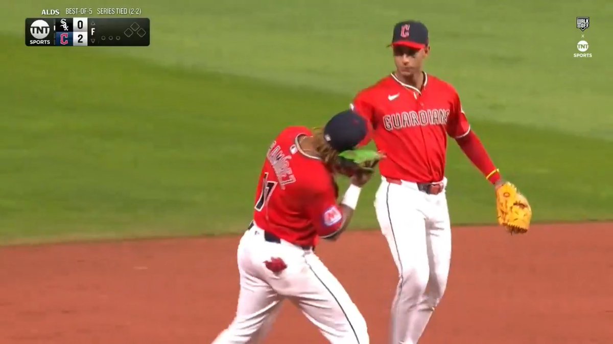 Two Cleveland Guardians players in red jerseys and caps celebrate on the field after their ALDS victory over the Chicago White Sox, with the scoreboard showing Cleveland leading 2-0 in a series tied 2-2 (best-of-5). The image captures the triumphant moment as the Guardians complete their comeback to advance to the ALCS.