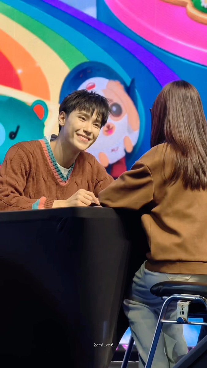 A young man in a brown sweater smiles while signing or drawing for a female fan in a brown jacket at what appears to be a fan event, with a colorful illustrated backdrop behind them.