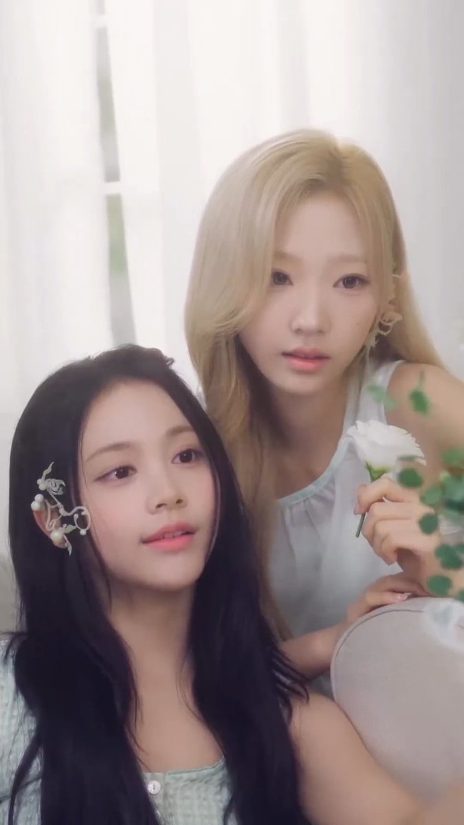 Two young women pose together in soft, hazy lighting; one has long dark hair with a delicate silver ear cuff, while the other has long blonde hair and holds a small white flower.