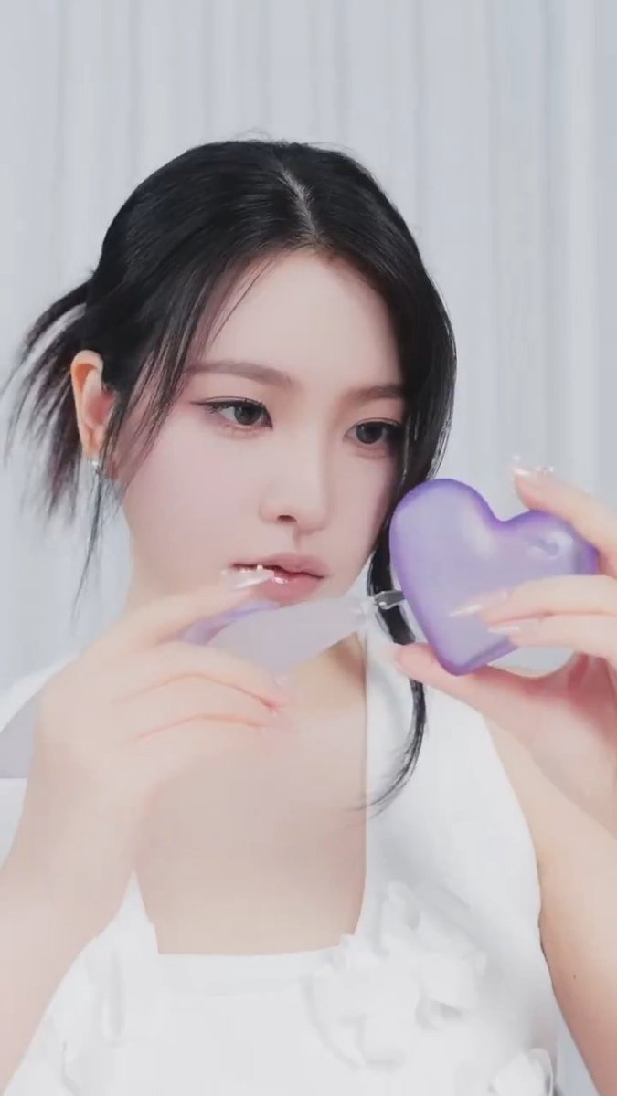 A close-up portrait of a woman with dark hair holding a purple heart-shaped object near her face, wearing a white top.
