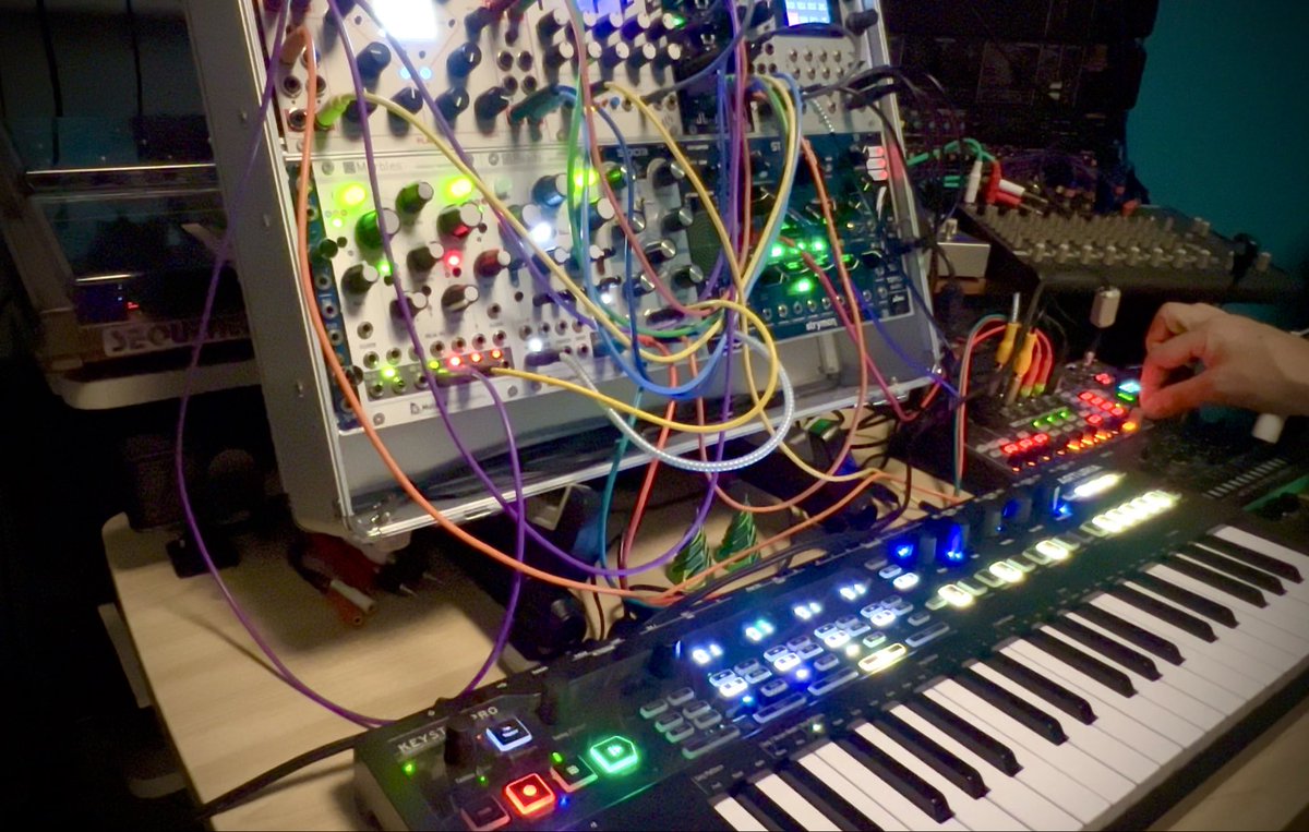 A complex modular synthesizer setup with colorful patch cables connecting various modules with illuminated LEDs, alongside a keyboard with glowing buttons, with a person's hand visible adjusting controls.
