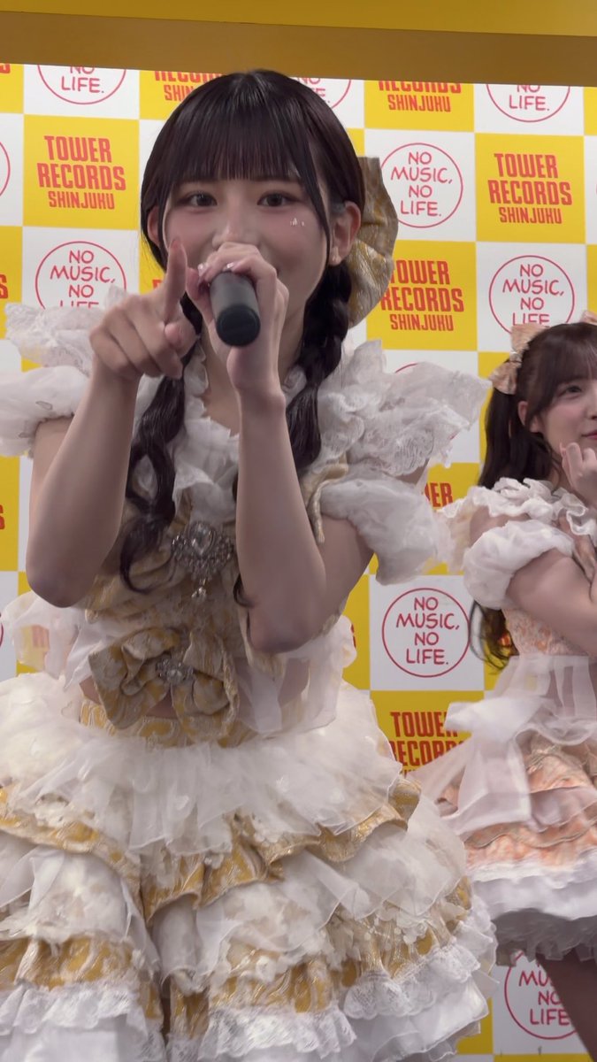 A young Japanese idol in an elaborate frilly white and gold dress speaks into a microphone while pointing upward. The backdrop features Tower Records branding with 'TOWER RECORDS SHINJUKU' and 'NO MUSIC, NO LIFE.' text.