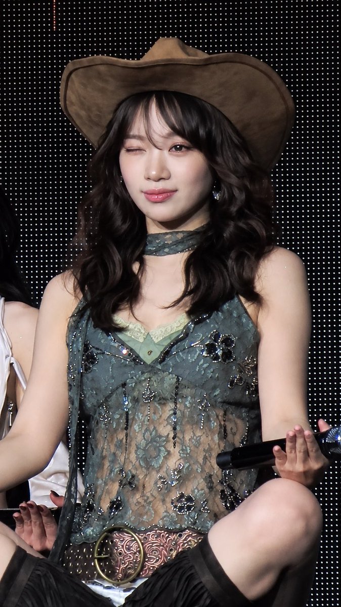 A young woman with long dark wavy hair winks while wearing a brown cowboy hat and a sheer, embellished green floral top with a choker, holding a microphone.