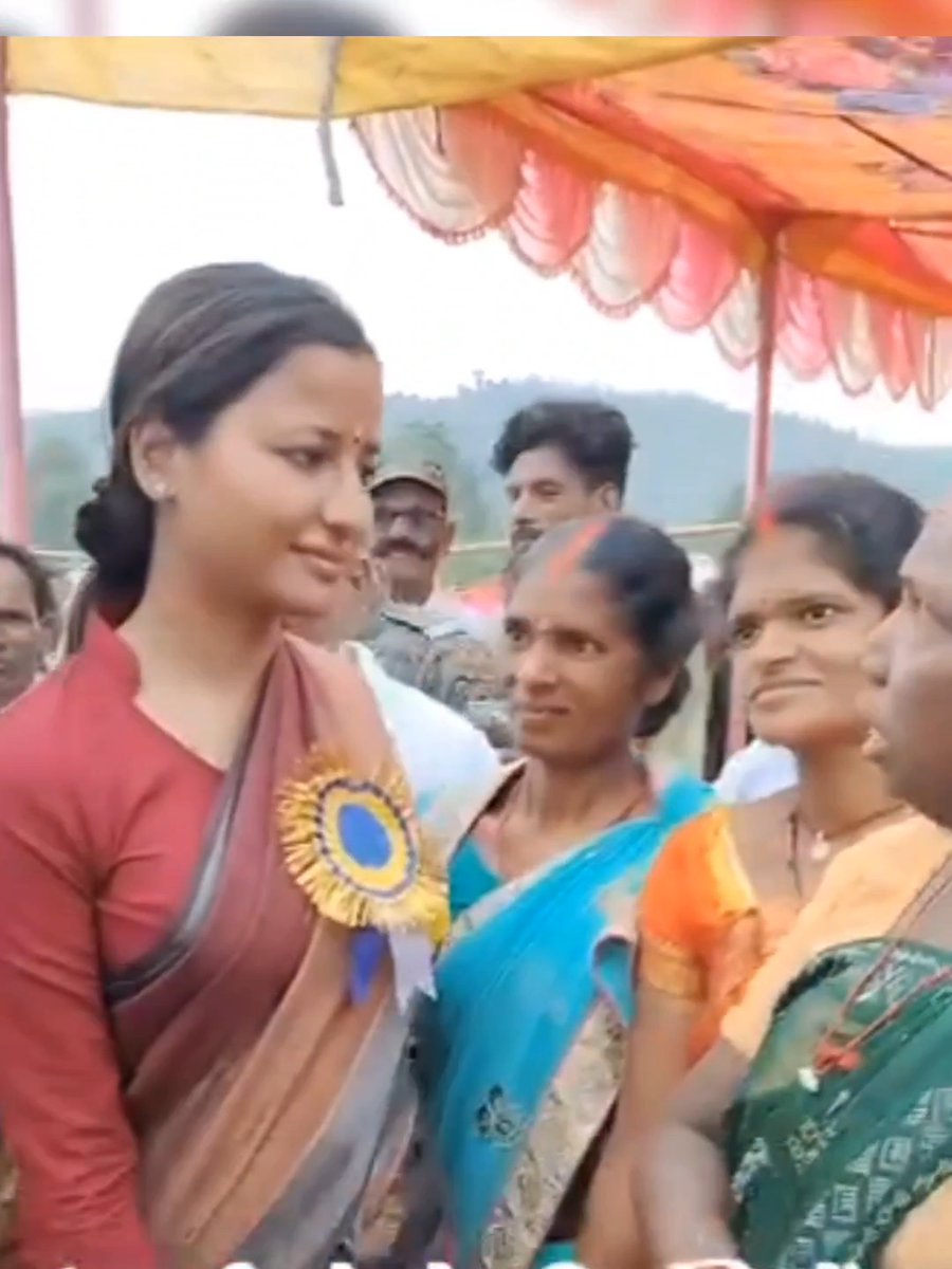 An outdoor event shows a woman in a maroon saree wearing a blue and yellow ribbon rosette, standing among other women in colorful traditional Indian attire under a tent.