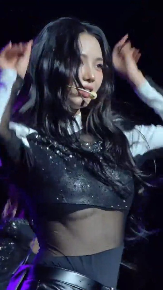 A young woman with long dark hair performing on stage, wearing a sparkly black crop top and a headset microphone, with her hands raised near her face.