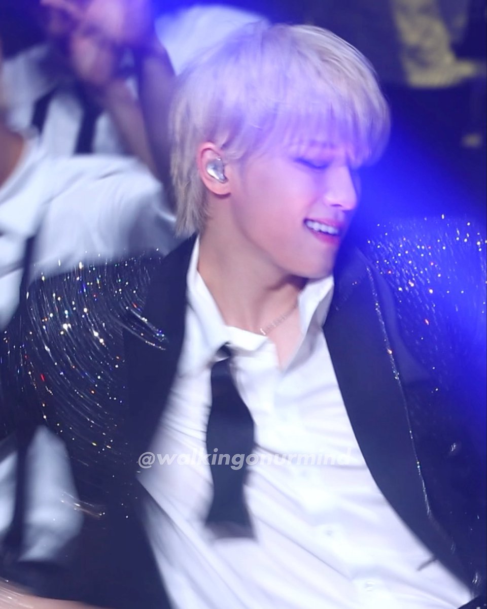 A performer with blonde hair on stage wearing a sparkly black jacket, white shirt, and black tie, with the watermark '@walkingonurmind_' visible.