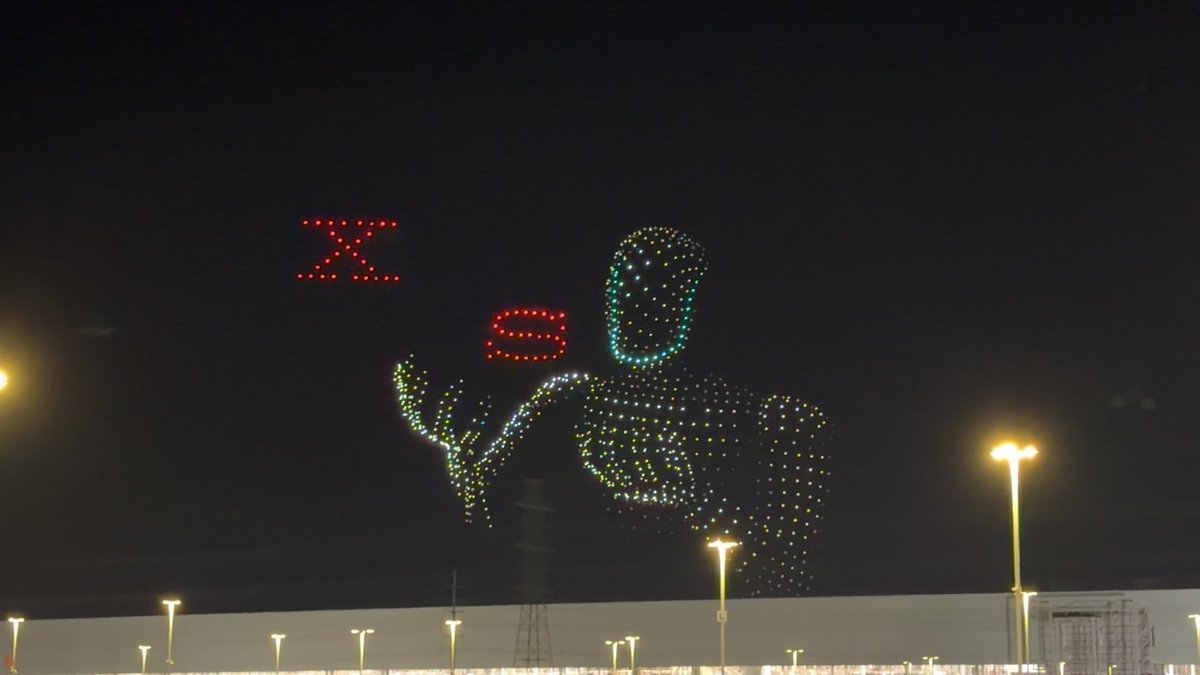 A nighttime drone light show displays a large humanoid figure and red Chinese characters in the sky above an illuminated parking lot.