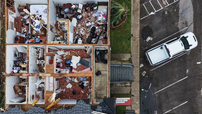 An aerial view shows a home with its roof torn off and interior contents scattered across rooms, with a damaged white vehicle visible in the adjacent parking lot. The destruction illustrates the aftermath of Hurricane Isaias, which killed at least four people after making landfall in Florida and left hundreds of thousands without power across the Southeast.
