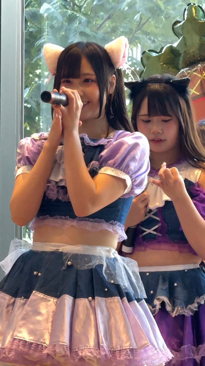 Two young women in purple and blue idol costumes performing, one with cat ears singing into a microphone while the other wears black cat ears.