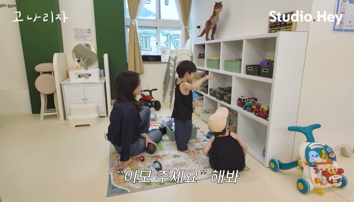 A still from a Korean TV show '고네리자' (Studio Hey) showing a woman sitting on a play mat with two young children in what appears to be a daycare or playroom with toy storage shelves.