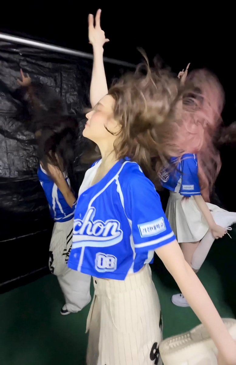 Two female performers in blue and white baseball-style uniforms dance energetically, with one prominently showing a jersey with "chon" and the number 08.