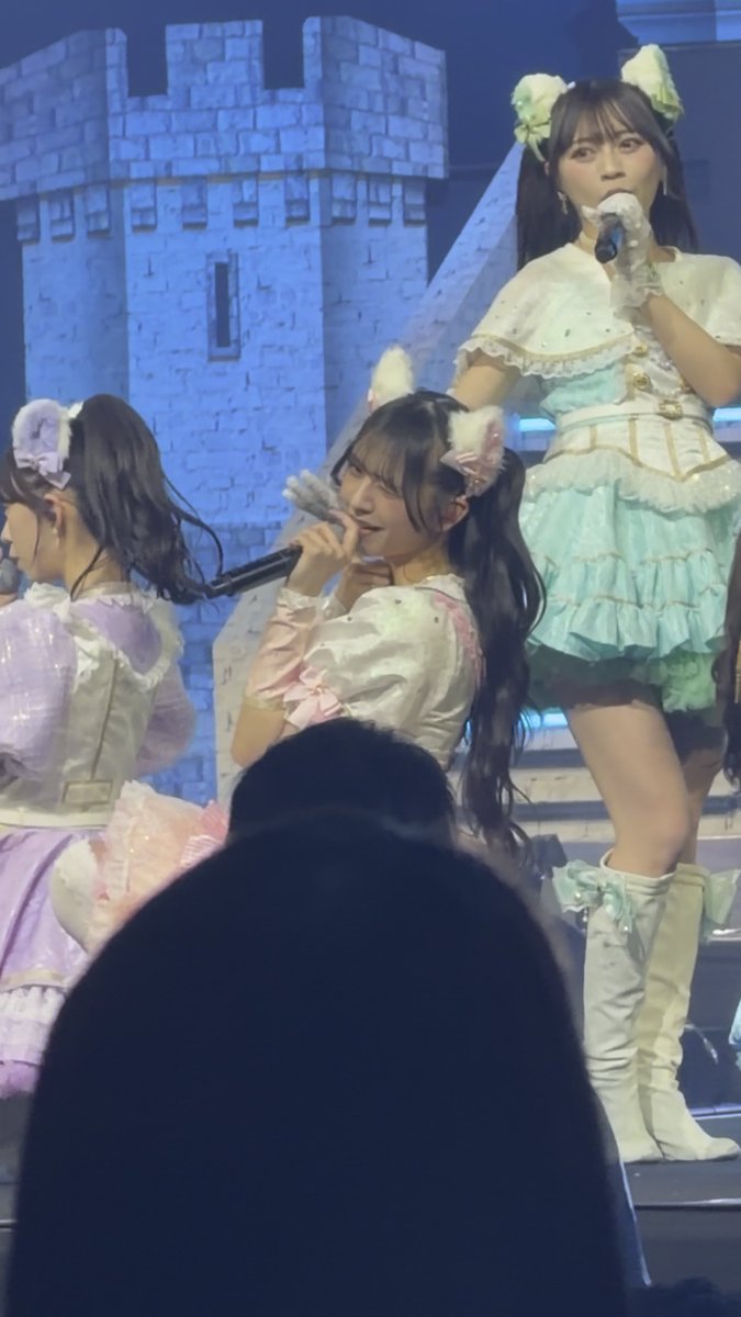 Three performers on stage in pastel-colored, frilly outfits with animal ear headbands, singing into microphones in front of a castle-themed backdrop.