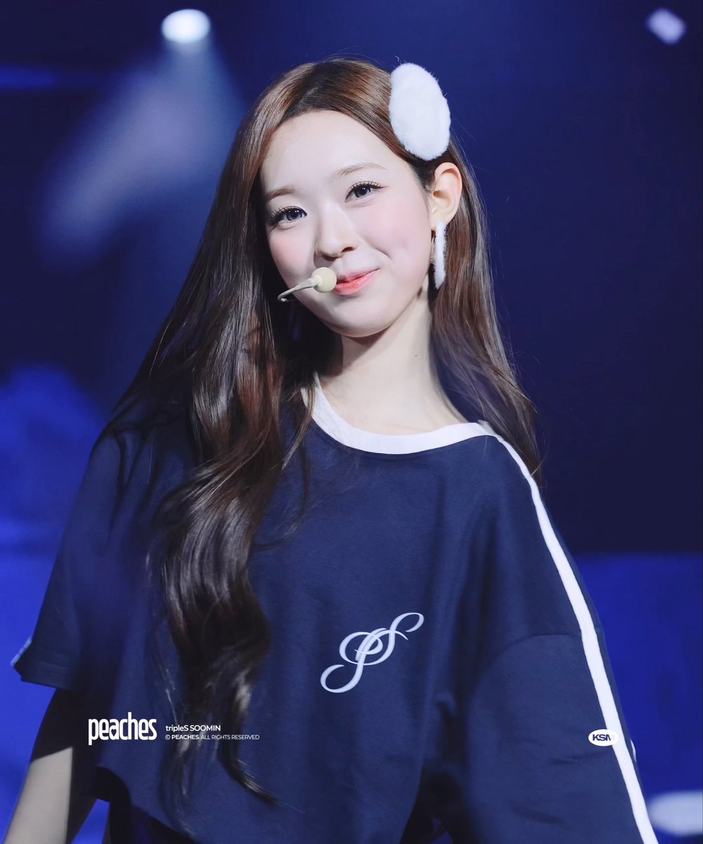 A young woman with long wavy brown hair and a white fuzzy hair accessory, wearing a blue t-shirt with a white logo and a headset microphone, performing on stage with a blue background. Text reads 'peaches tripleS SOOMIN'.