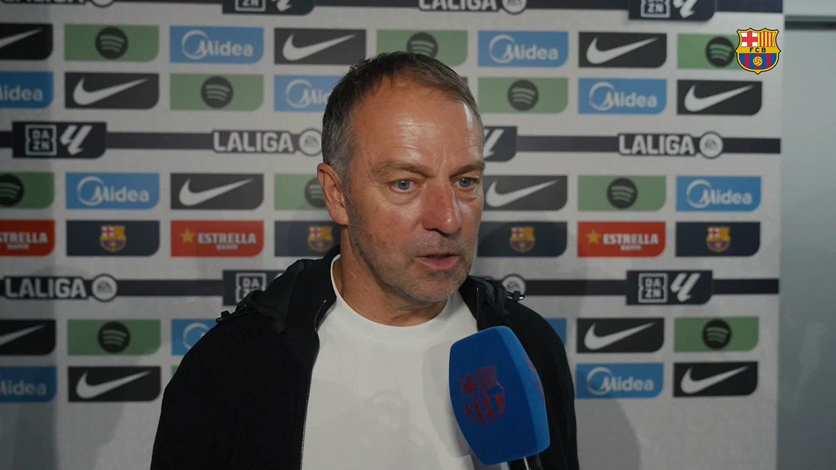 Hansi Flick speaks into a microphone during a post-match interview after FC Barcelona's 3-0 La Liga victory over Getafe, with a backdrop featuring sponsor logos including Midea, Nike, Estrella, and the FCB crest.