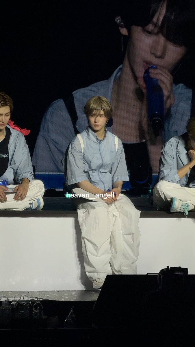 A performer with blonde hair sits on a stage wearing a light blue shirt and white pants, with a giant screen behind him showing him singing into a blue microphone; the watermark reads "heaven_angell".