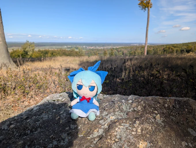 Cirno fumo doll sitting on a stone in a forest, sky and city in the background