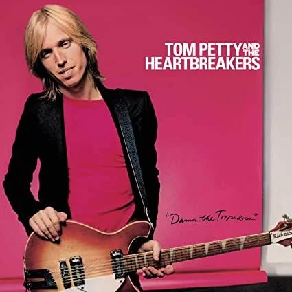 Album cover of Tom Petty and the Heartbreakers' Damn the Torpedoes: Tom Petty, with long blond hair, in a black jacket over a pink T-shirt, holding a sunburst Rickenbacker guitar against a hot-pink backdrop. "TOM PETTY AND THE HEARTBREAKERS" is printed in white at the top right, with "Damn the Torpedoes" handwritten below.