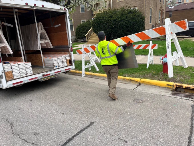 Staff in a yellow safety vest carrying a long orange and white striped barricade to the curb.