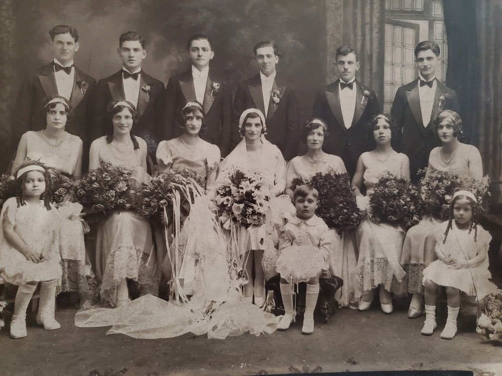 An old sepia-toned formal group portrait, likely of a wedding party. Men in suits stand behind women in long dresses holding bouquets, with several children in front. There is no visible text.